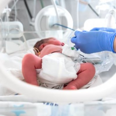 A pre-term baby lies in a humidi-crib while two blue gloved hands administer medicine. 